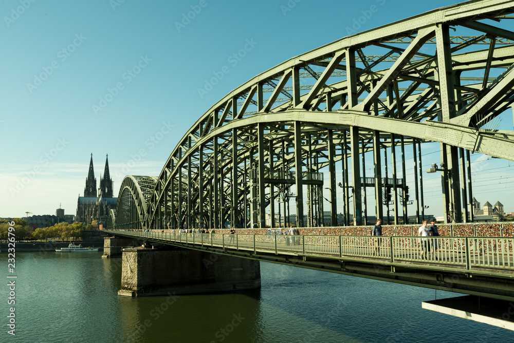 he Cologne Cathedral with Hohenzollern Bridge in the foreground on a bright sunny day. People are walking on the bridge he Cologne Cathedral with Hohenzollern Bridge in the foreground on a bright sunny day. People are walking on the bridge