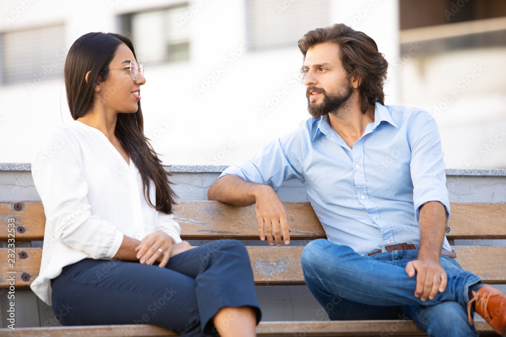 Successful multicultural couple talking while sitting on bench in city. Professional executive couple on meeting outdoors. Teamwork concept Successful multicultural couple talking while sitting on bench in city. Professional executive couple on meeting outdoors. Teamwork concept