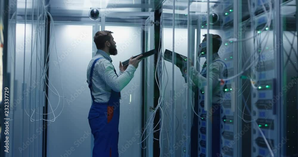 Medium shot of a male technician working in a data center carrying peforming maintenance amongst rows of server racks installing server equipment Medium shot of a male technician working in a data center carrying peforming maintenance amongst rows of server racks installing server equipment