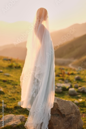 fine-art wedding. Beautiful girl with gorgeous slim in gray dress with hand-painted flowers stands against the mountain landscape at sunset
