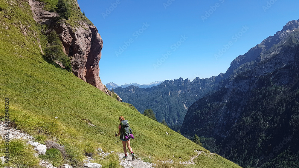 Hiker with backpack walking a grass trail on top of a mountain and enjoying valley view during sunny trip in the alps