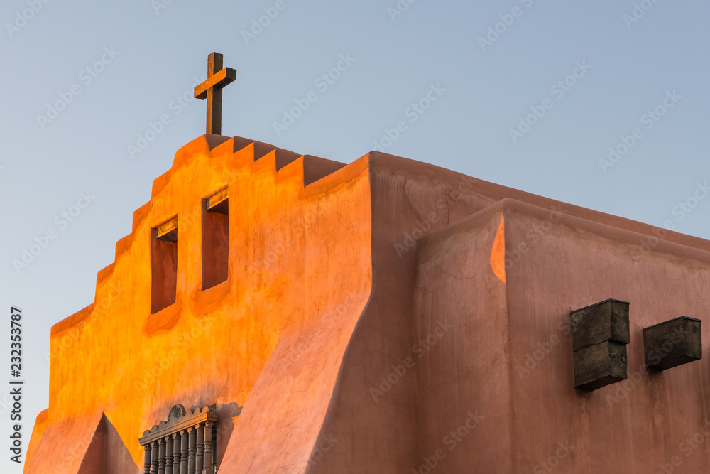 Fototapeta premium Kościół Adobe i rustykalny drewniany krzyż świecą w złotym wieczornym świetle w Santa Fe w Nowym Meksyku