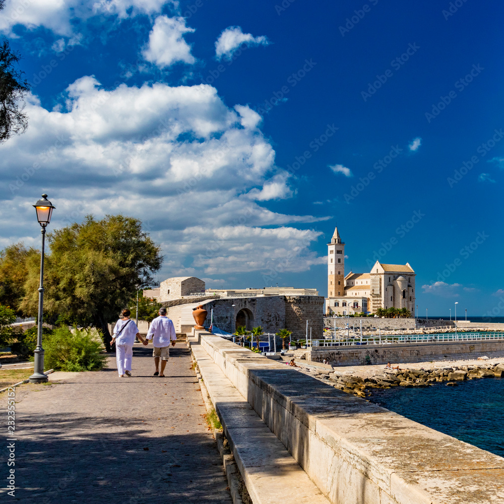 Two lovers walk, holding hands, in the municipal park of Trani, with the view of the sea and the Cathedral Basilica of St. Nicholas Pilgrim in the background. In Puglia, near Bari, Barletta, Andria.