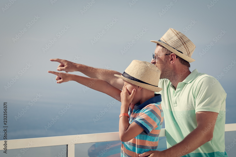 Dad and son staying at the hotel’s balcony against the ocean and pointing on something ahead.