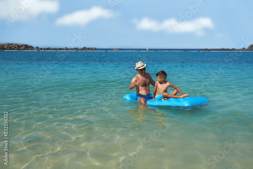 Dad and son are sitting on a blue inflatable floater in the clear ocean water. They are looking to each other, smiling, having fun and enjoying their vacations.