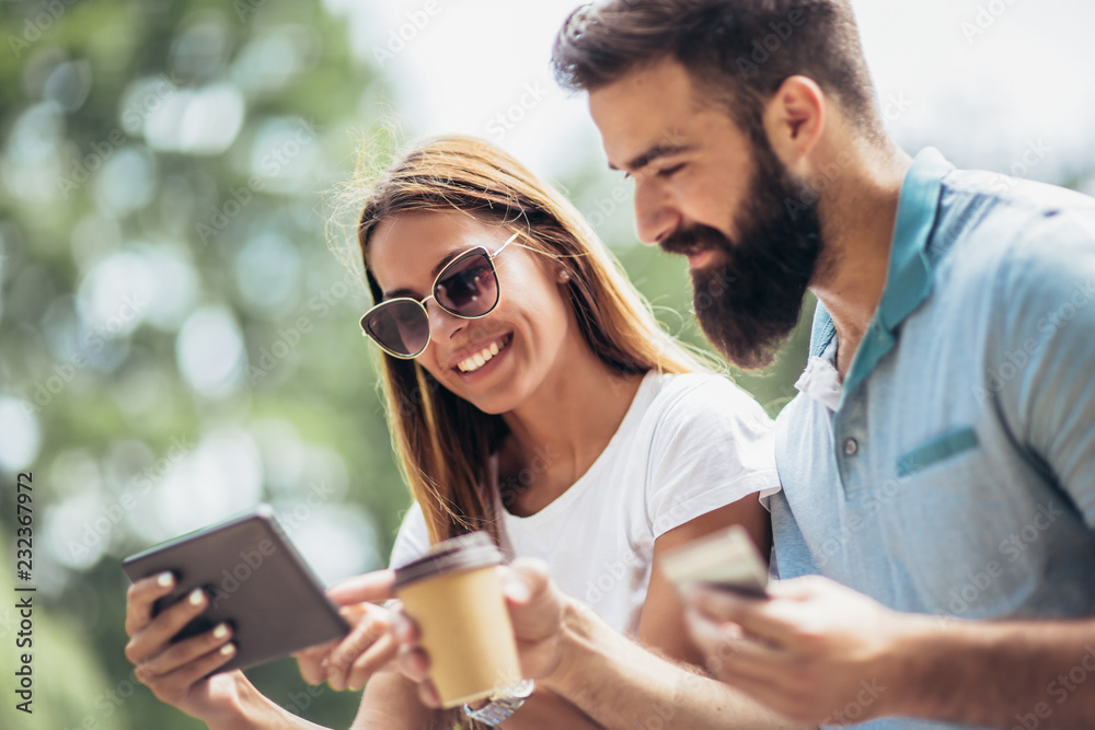 Cheerful young couple sitting on a park bench and uses a digital tablet for online shopping Cheerful young couple sitting on a park bench and uses a digital tablet for online shopping