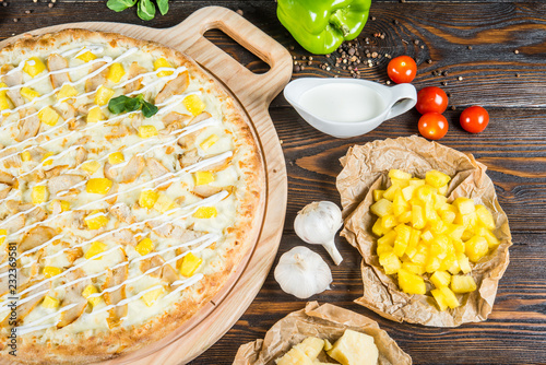 Large creamy pizza with white sauce, chicken and pineapples on a round cutting board on a dark wooden background. Ingredients.