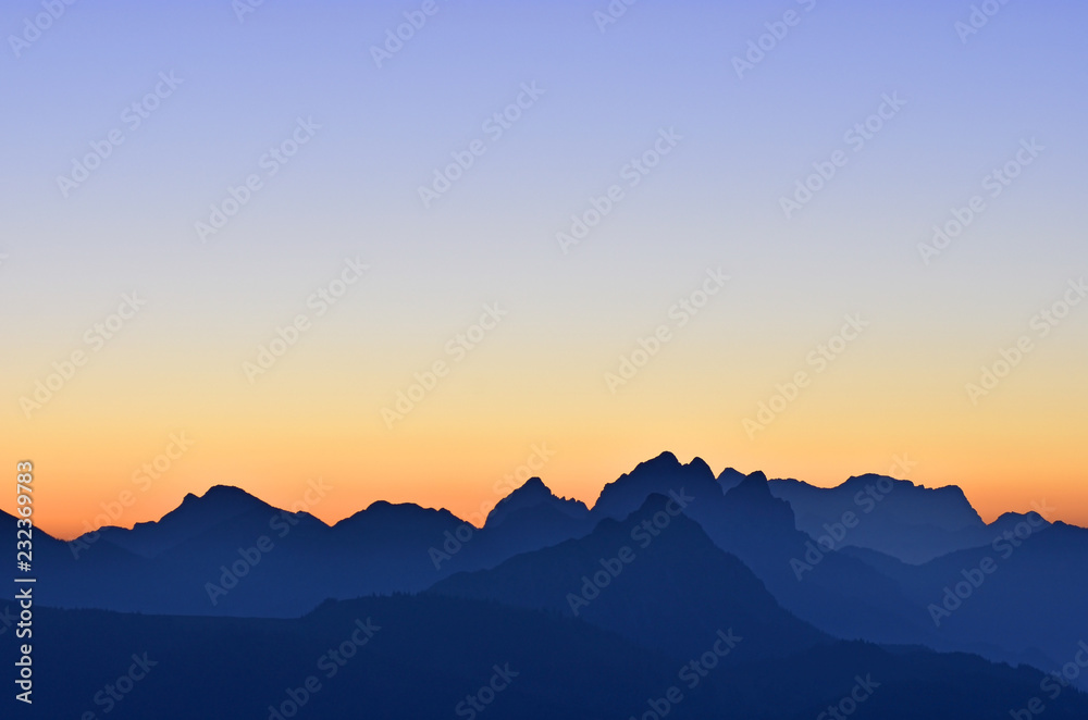 Beautiful morning mood in the Allgaeu Alps at the border region of Germany and Austria. Blue silhouettes of mountain ranges at dawn with orange and blue sky. Zugspitze in the background. Copy space.