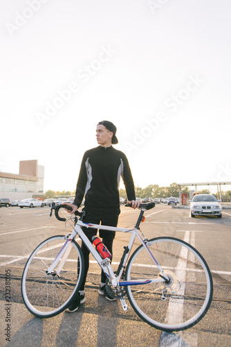 Young man is a cyclist with dark sportswear and a cap with a bicycle on the backdrop of the city landscape and looking to the side. Cycling Sports Concept.