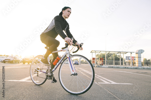 Fototapeta Naklejka Na Ścianę i Meble -  Portrait of an attractive cyclist in motion. Young rider in dark bike wears a bike on a street background and sunshine in the sunset.