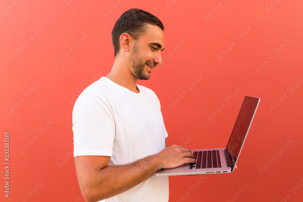 Handsome man in casual t-shirt holding silver notebook and chatting or working isolated over red background Handsome man in casual t-shirt holding silver notebook and chatting or working isolated over red background