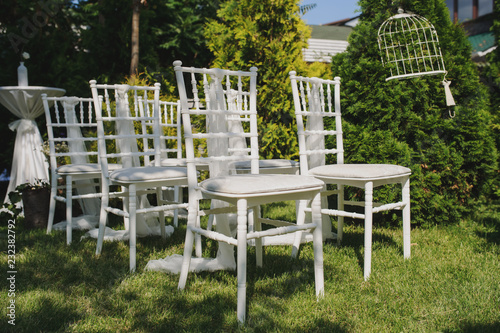 Rows of white empty chairs on a lawn before a wedding ceremony sorrunded by trees with white vintage cages on them and with a catering white table with a cage on it