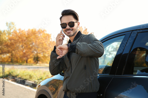 Young man checking time while talking on phone near modern car, outdoors