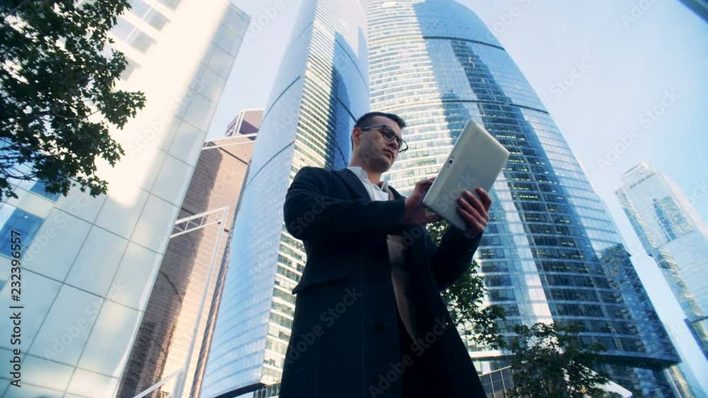 Successful businessman is operating his tablet while standing next to skyscrapers. 6K Cinema camera footage.