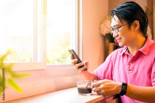 Asian man or entrepreneur holding a cup of coffee using smartphone to surf internet or checking stock from their shop during relax time in the morning at home or coffee shop. Business and Technology.