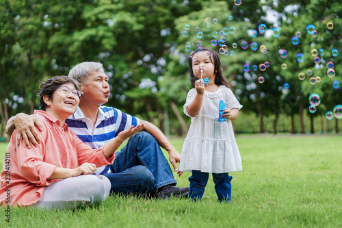 Asian cute granddaughter making bubbles and attractive happy grandparents look at her in the public garden at vacation day or holiday. Family Concept. photo