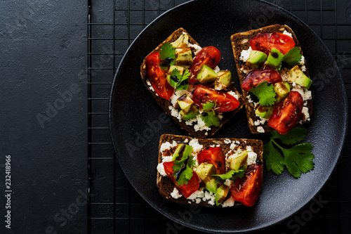 Avocados, cherry tomatoes and feta cheese sandwiches with balsamic sauce and cilantro on old olive board on dark black background. Top view.