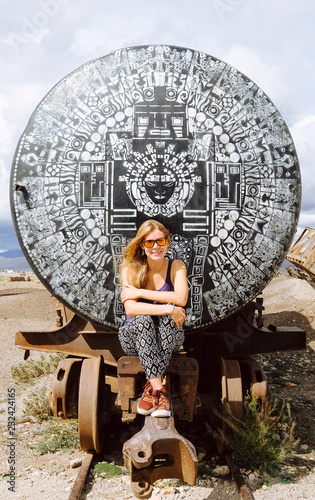 Bolivia. Cemetery of old trains. The blonde girl sits on a train car in sunglasses. Clear day. The train car is painted with an unusual black-and-white pattern in a mazaic style. photo