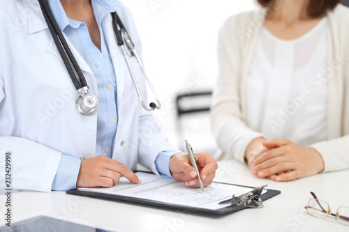Unknown doctor and female patient discussing something while standing near reception desk in emergency hospital. Physician at work in clinic. Medicine and health care concept