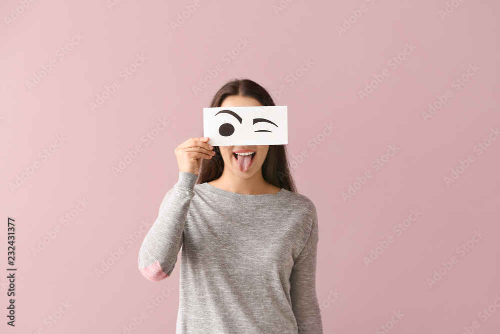 Emotional young woman hiding face behind sheet of paper with drawn eyes on color background Emotional young woman hiding face behind sheet of paper with drawn eyes on color background