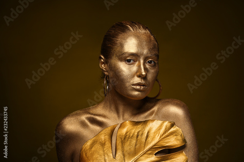 Beautiful young woman with golden paint on her body and tropical leaf against dark background