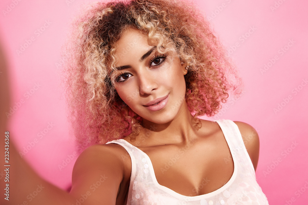 Portrait of blissful lovable woman with blond african hairstyle. Smiling model girl in trendy summer clothes taking selfie on her smartphone for social media. Posing on pink background