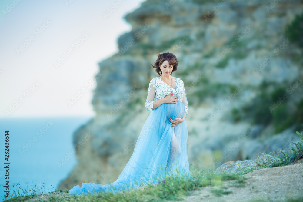 Young attractive pregnant lady woman wearing casual blue airy dress posing close to ocean sea cape mountains over sky background Young attractive pregnant lady woman wearing casual blue airy dress posing close to ocean sea cape mountains over sky background