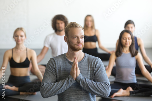 Young handsome male instructor and a group of young sporty people practicing yoga lesson, doing Padmasana exercise, Lotus pose, working out, indoor close up portrait photo, studio, namaste gesture