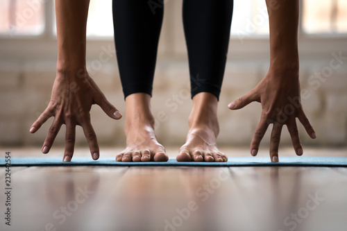 Sporty active woman practicing yoga, doing Standing forward bend exercise, head to knees uttanasana pose, wearing black sportswear pants, indoor close up, yoga studio or home. Well being concept