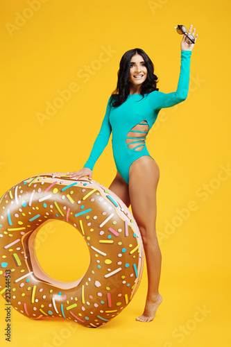 Full lenhts portrait of an excited young woman dressed in swimsuit holding sunglasses and posing with inflatable ring on the orange background.