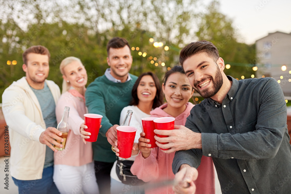 Fototapeta premium leisure and people concept - happy friends with drinks taking selfie at rooftop party in summer