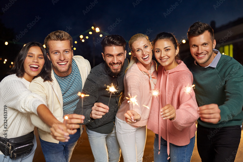 Obraz premium leisure, celebration and people concept - happy friends with sparklers at rooftop party at night