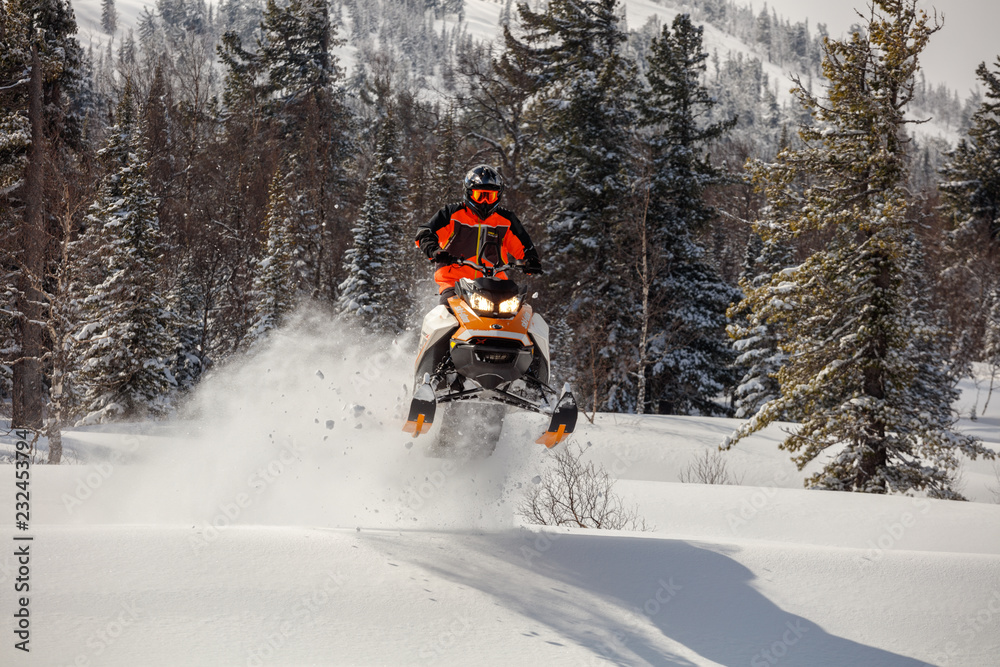 the guy is flying and jumping on a snowmobile on a background of winter forest  leaving a trail of splashes of white snow. bright snowmobile and suit without brands. extra high quality