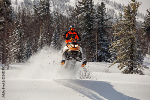 the guy is flying and jumping on a snowmobile on a background of winter forest leaving a trail of splashes of white snow. bright snowmobile and suit without brands. extra high quality