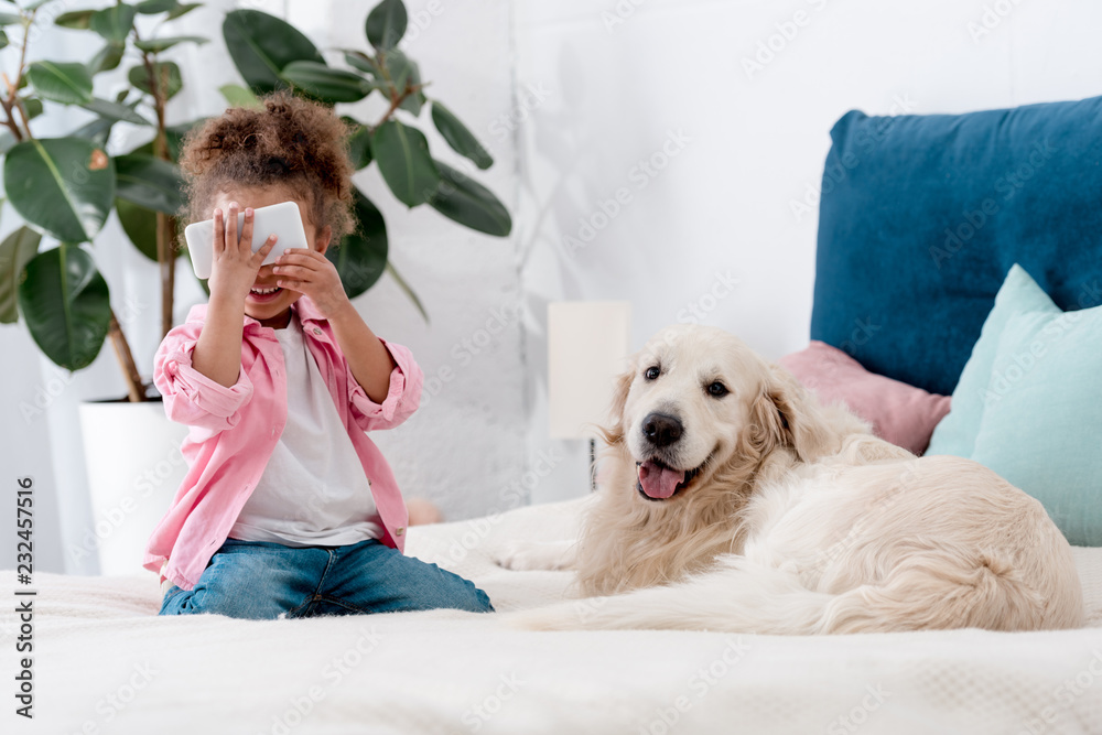 cute african american child sitting on the bed with retriever and holding smartphone near face cute african american child sitting on the bed with retriever and holding smartphone near face