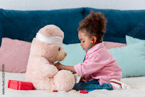 Little african american child in pink jacket playing the doctor  with teddy bear