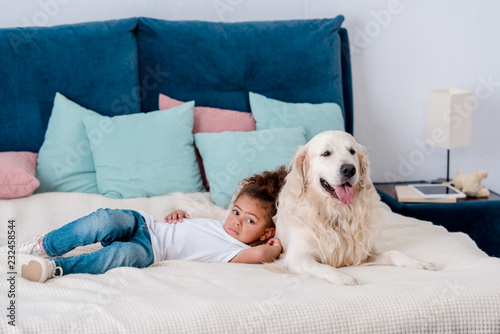 Adorable little african american kid lying on bed and leaning to happy dog