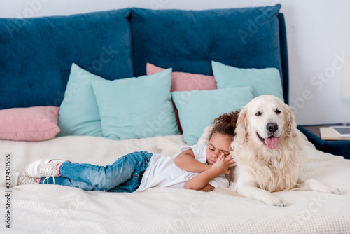 Cute little african american kid lying on bed and leaning to dog