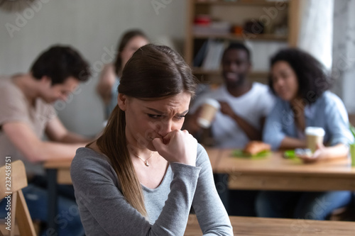 Diverse multi-ethnic friends sitting together in cafe talking having fun, focus on frustrated shy girl sitting separately by others teenagers feels unhappy because peers not accept her she is outcast