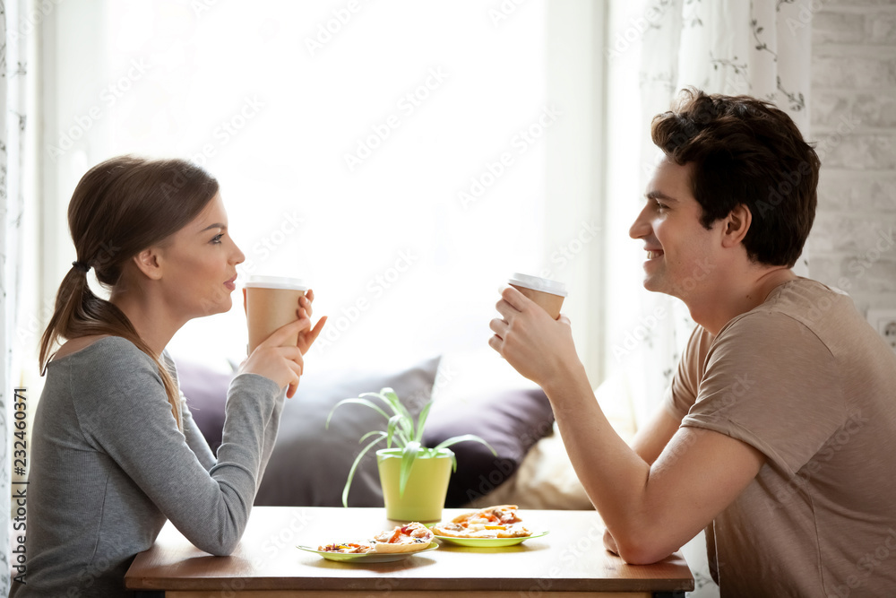 Happy positive couple in love attractive girlfriend handsome boyfriend looking at each other drinking coffee from paper cups enjoy time together sitting at desk near window in cozy pizzeria, side view Happy positive couple in love attractive girlfriend handsome boyfriend looking at each other drinking coffee from paper cups enjoy time together sitting at desk near window in cozy pizzeria, side view