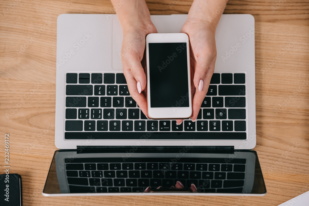 cropped shot of woman holding smartphone in hands at tabletop with laptop