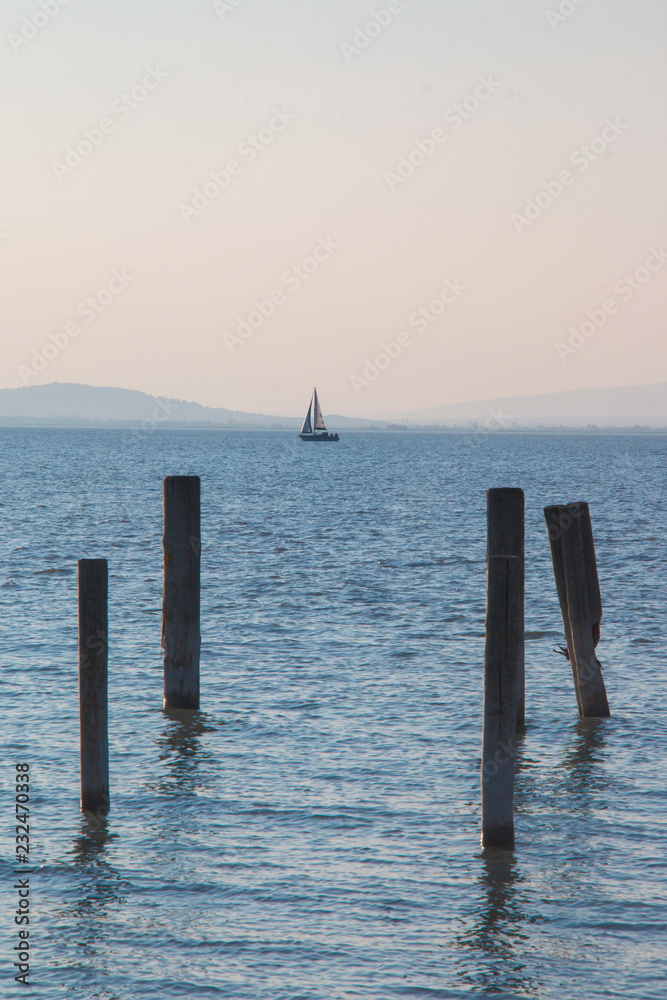 Wooden dock pillars vertically in light blue sea water and small holiday sailboat sailing in distance on horizon, idea of vacation activity on sea Wooden dock pillars vertically in light blue sea water and small holiday sailboat sailing in distance on horizon, idea of vacation activity on sea