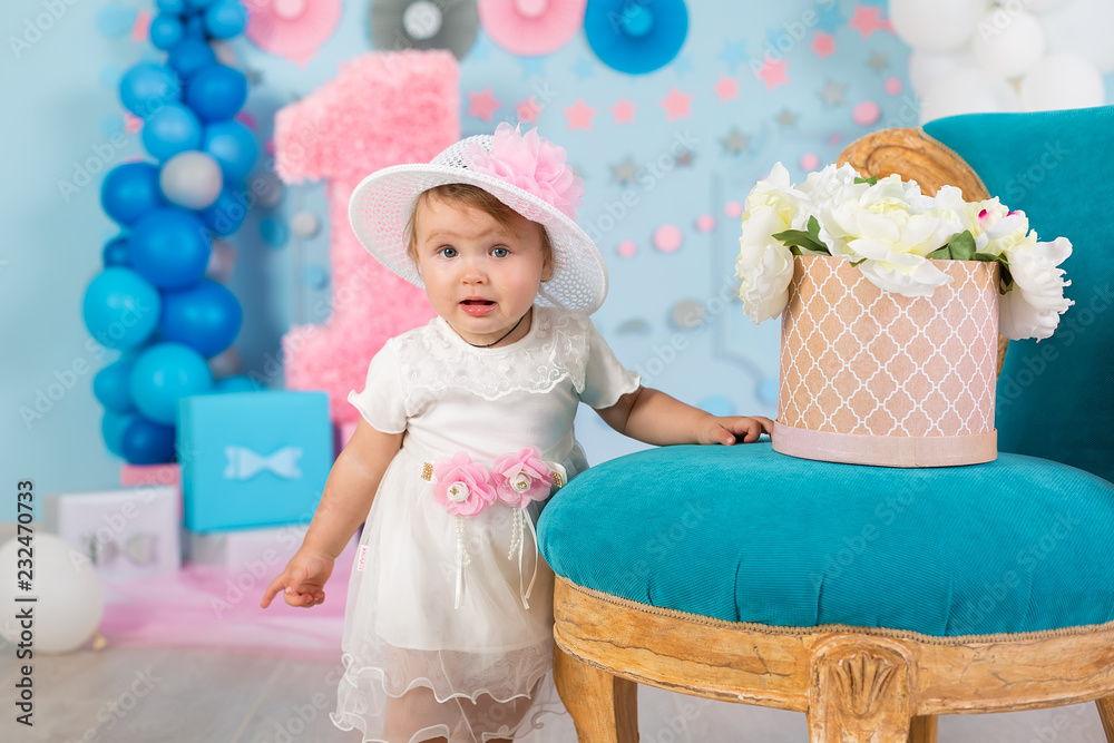 Fototapeta premium Cute little baby girl with big blue eyes wearing tutu hat and flower in her hair posing sitting in studio decorations with number one celebrating her birthday