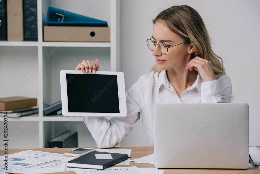 portrait of businesswoman in eyeglasses showing tablet with blank screen at workplace with papers and laptop in office portrait of businesswoman in eyeglasses showing tablet with blank screen at workplace with papers and laptop in office