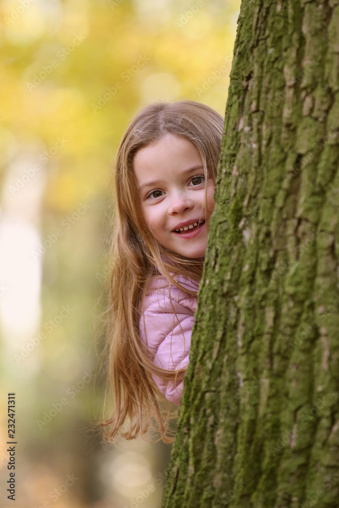 Fototapeta premium Kleines Mädchen schaut lachend hinter einem Baum hervor