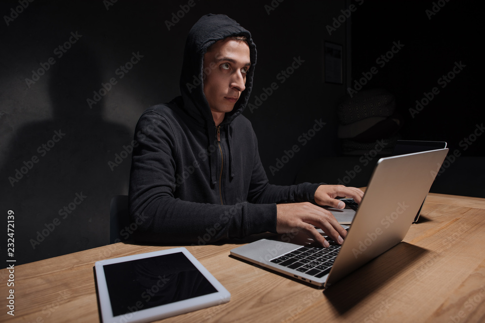 hacker in black hoodie using laptops at tabletop with tablet in dark room, cyber security concept hacker in black hoodie using laptops at tabletop with tablet in dark room, cyber security concept