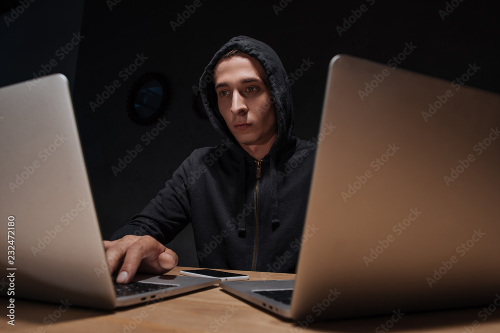 portrait of hacker in black hoodie using laptops at tabletop with smartphone, cyber security concept