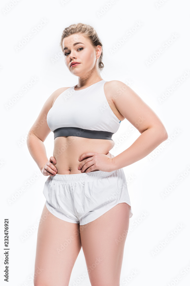low angle view of young overweight woman in sportswear standing with hands on waist and looking at camera isolated on white low angle view of young overweight woman in sportswear standing with hands on waist and looking at camera isolated on white