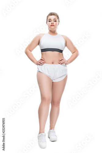 full length view of young size plus woman in sportswear standing with hands on waist and looking at camera isolated on white photo