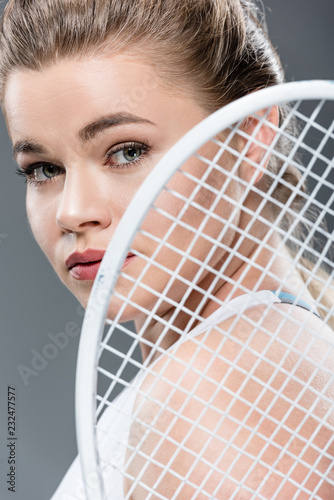close-up view of young woman holding tennis racket and looking at camera isolated on grey photo